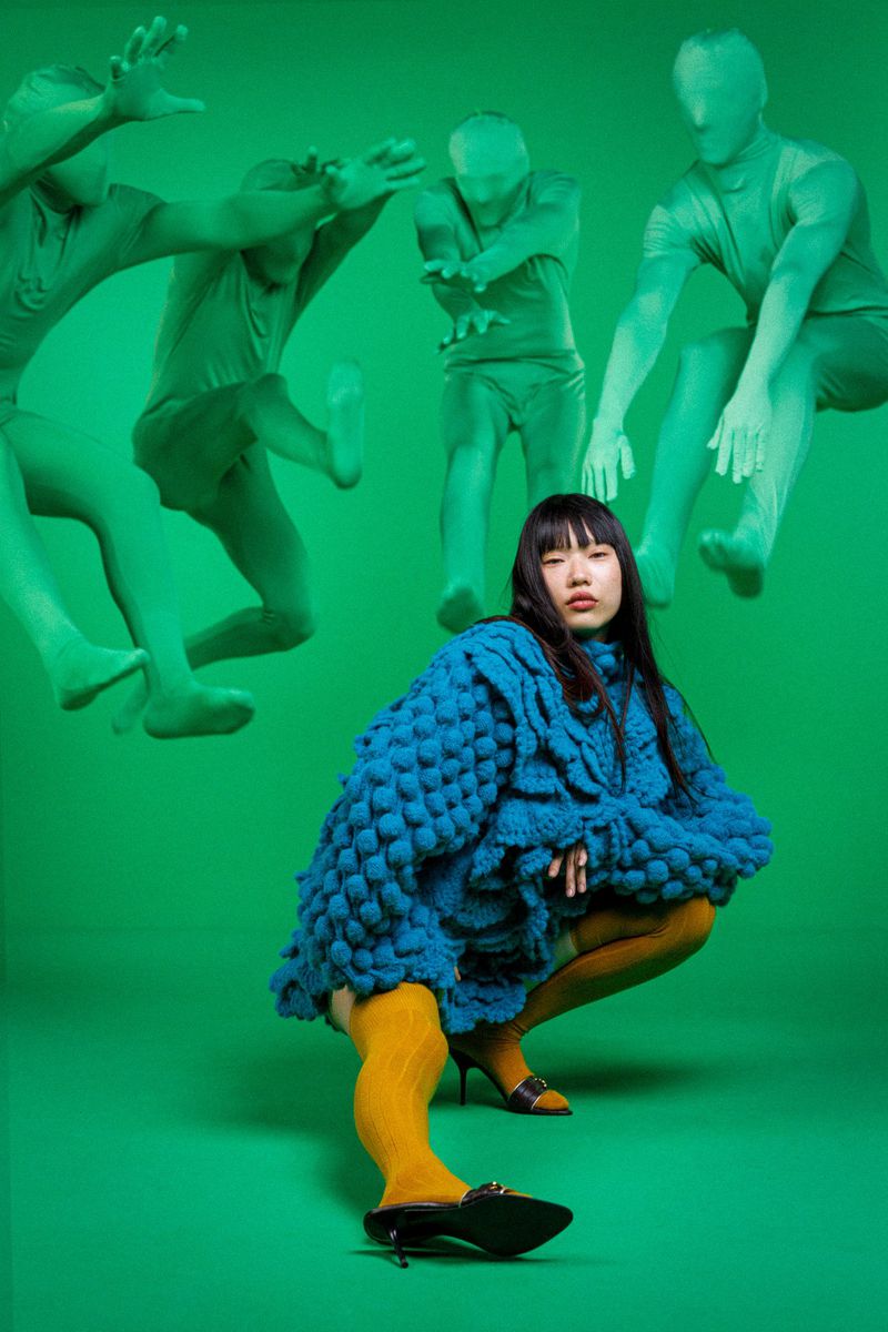 A young Asian woman poses in a blue sweater with a green background and blurred figures, wearing yellow tights and black heels.