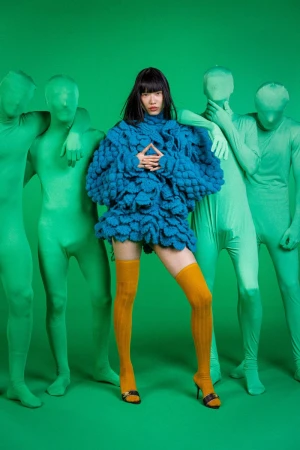 Asian woman wearing a knit dress and mustard stockings poses in front of four figures wearing green full body suits in a studio.