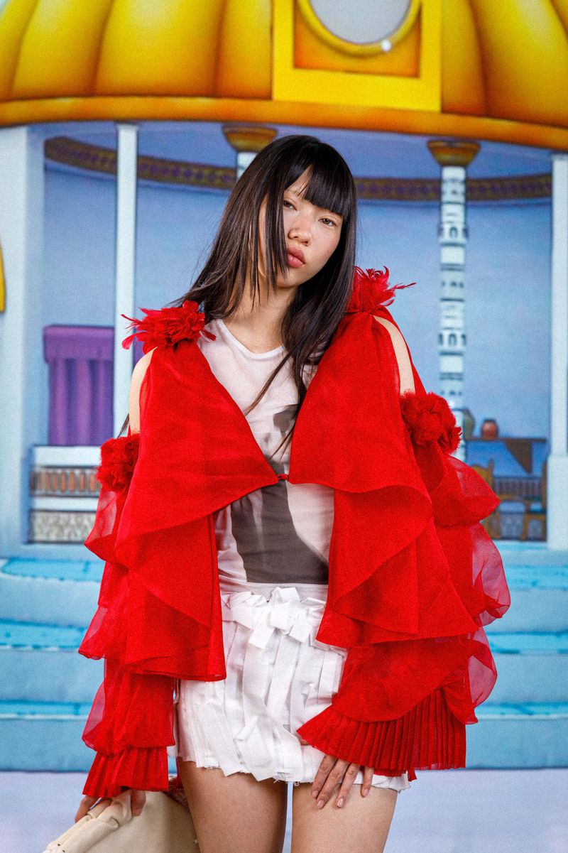 Woman in red ruffled jacket and white shorts poses in front of a blue and yellow backdrop, looking at the camera in a fashion shot.
