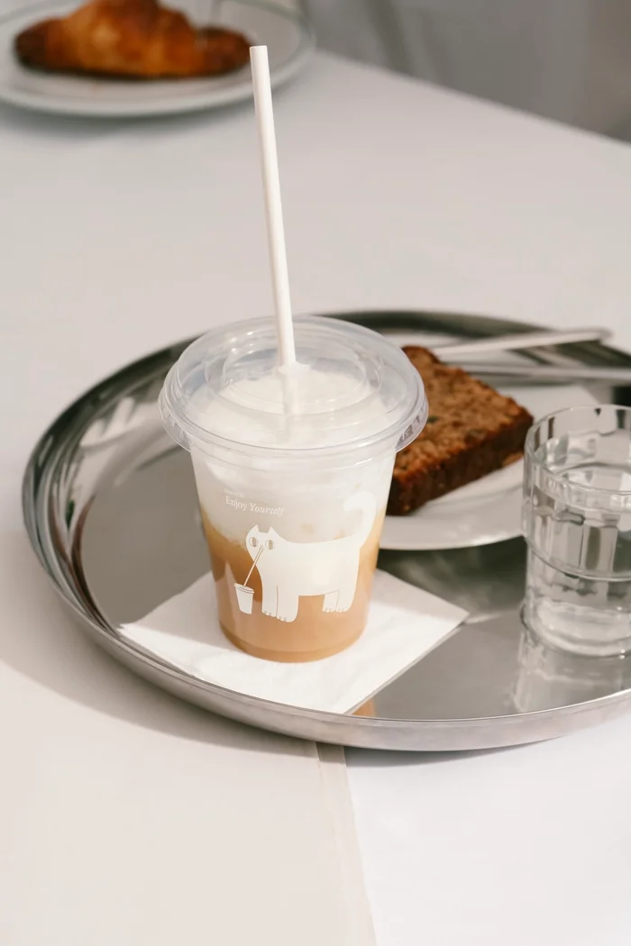 Iced coffee in a clear plastic cup with white straw on a metal tray with a slice of cake