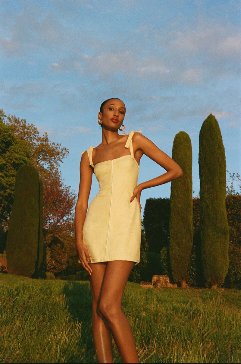 An adult female poses in a pale yellow mini dress in a grassy outdoor environment with cypress trees under a blue sky, fashion shot.
