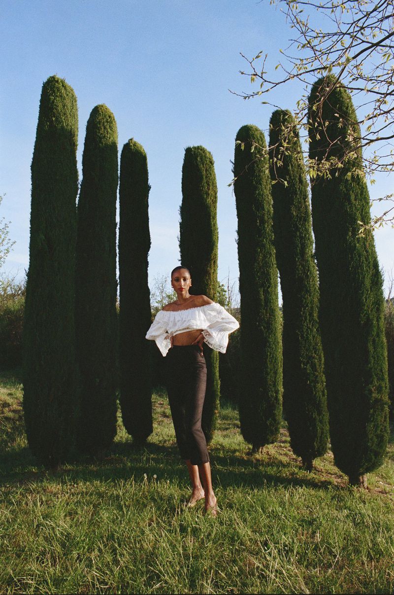 Woman in off-shoulder top and dark skirt stands in grassy field with tall, columnar trees under a bright blue sky, bathed in sunlight.