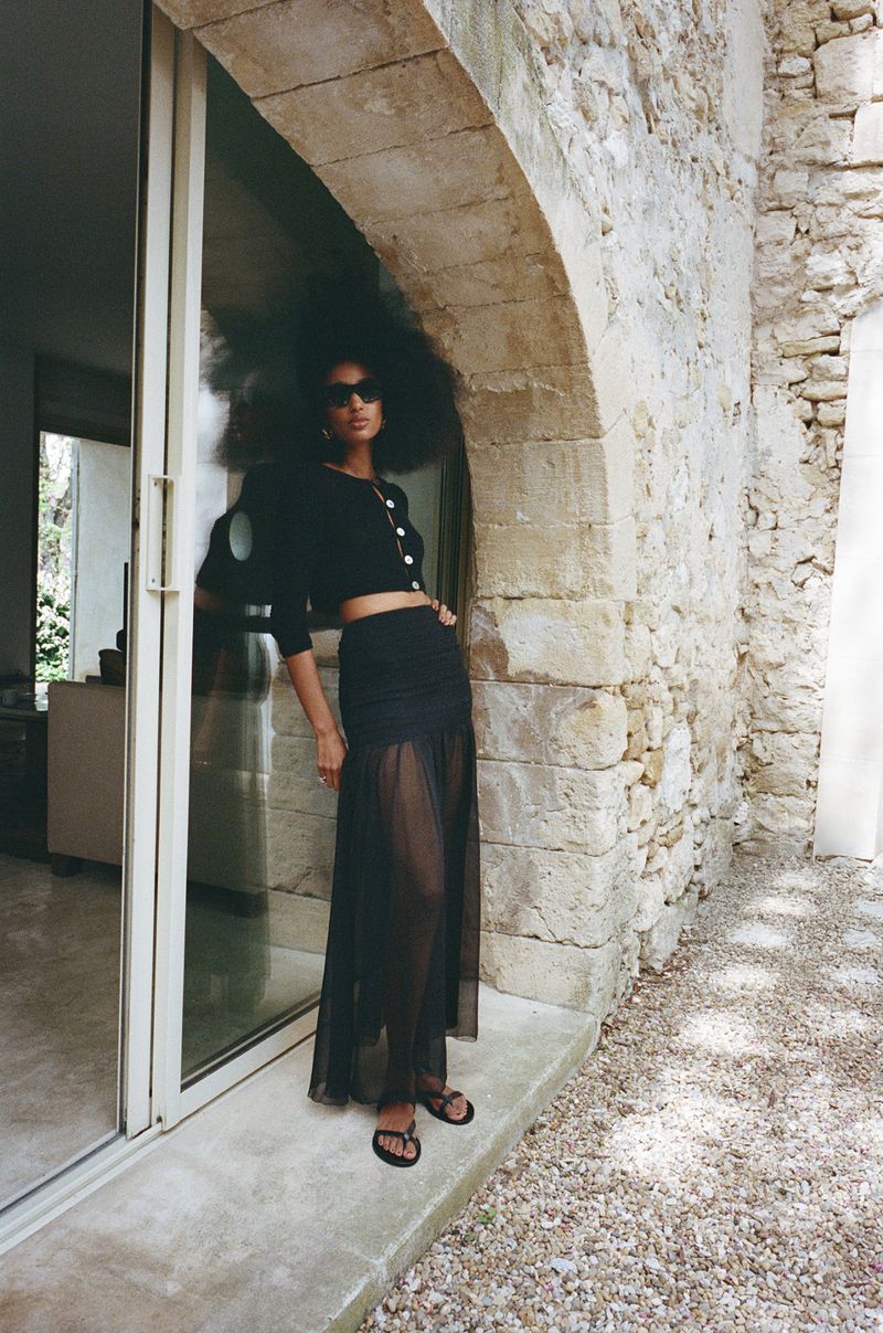 An adult female model poses next to an open doorway with an arch in a black, semi-sheer maxi skirt, cropped sweater, and sandals.