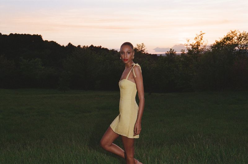 An adult female model poses in a yellow dress in a grassy field during golden hour, with a treeline and sky in the background.
