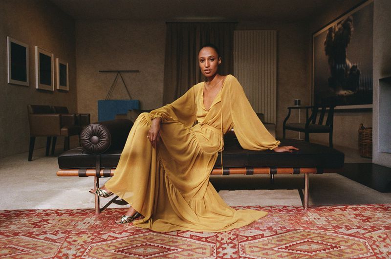 A woman in a yellow dress sits on a leather daybed on a red patterned rug in a room with neutral colors and modern art.