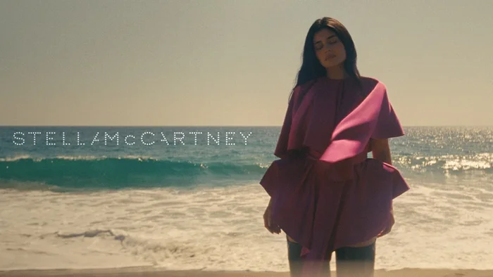 Kylie Jenner wearing a magenta cape top standing on a sandy beach by the ocean for Stella McCartney Wild Horses