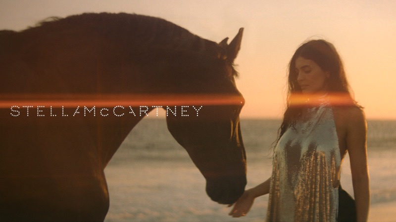 Kylie Jenner standing on a sandy beach next to a dark horse at sunset for Stella McCartney