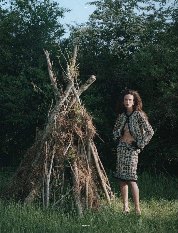 Model Waleska Gorczevski in a tweed jacket standing near a rustic wooden structure outdoors in a grassy field