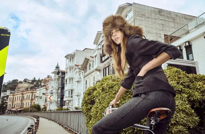 Model wearing a furry winter hat and dark grey outfit riding a bicycle along a waterfront street with traditional buildings.