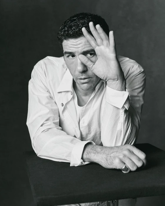 Monochrome portrait of actor Tahar Rahim in a white shirt, holding his hand up near his face, leaning on a dark table.