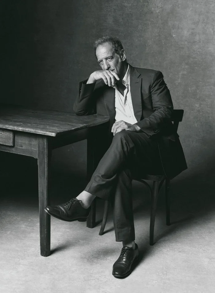 Monochrome portrait of a middle-aged man seated at a wooden table in a studio setting by Zeb Daemen for Vanity Fair France.