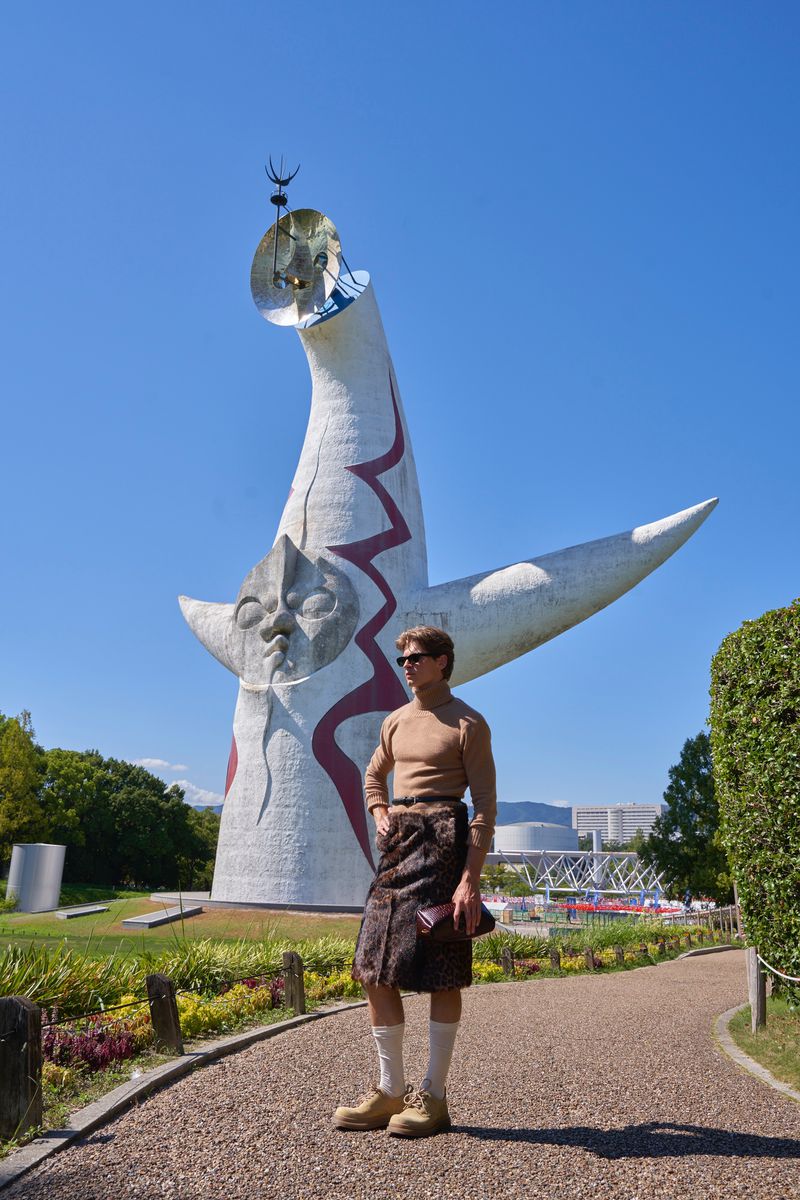Model wearing a tan turtleneck and leopard print skirt posing on a gravel path before a large sculptural monument