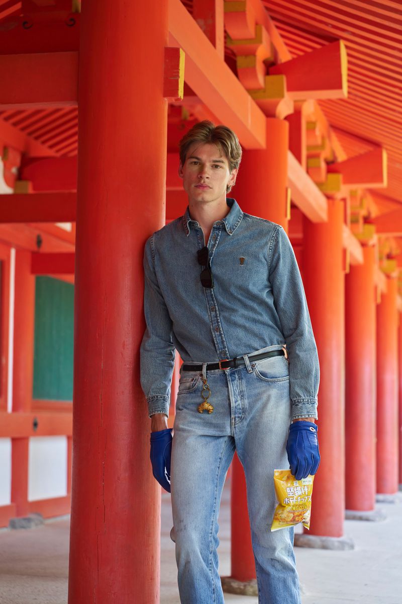 Model leaning against a bright red wooden pillar wearing a denim shirt, light wash jeans, and blue gloves holding a chip bag