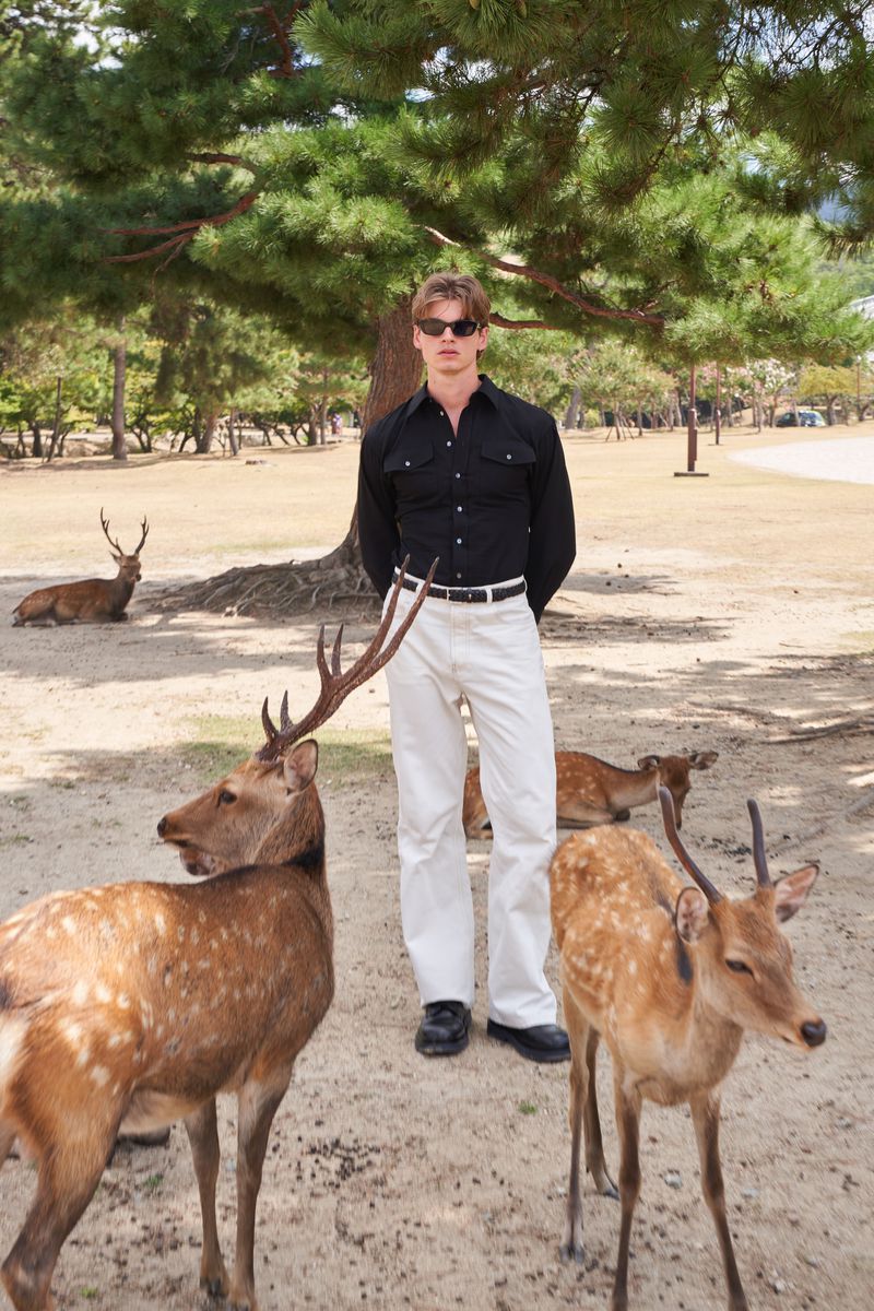 Male model in black shirt and white trousers standing outdoors among several deer in a park setting.