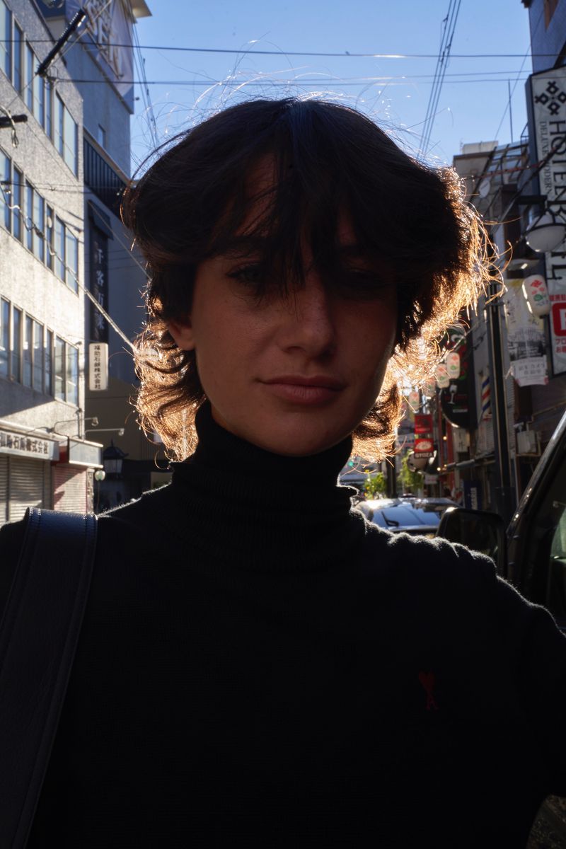 Model with dark hair wearing a black turtleneck sweater standing on a city street.