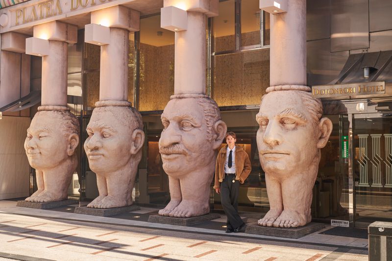 Model standing next to giant stone head columns outside Dotonbori Hotel for ODDA Magazine editorial by Regine David.