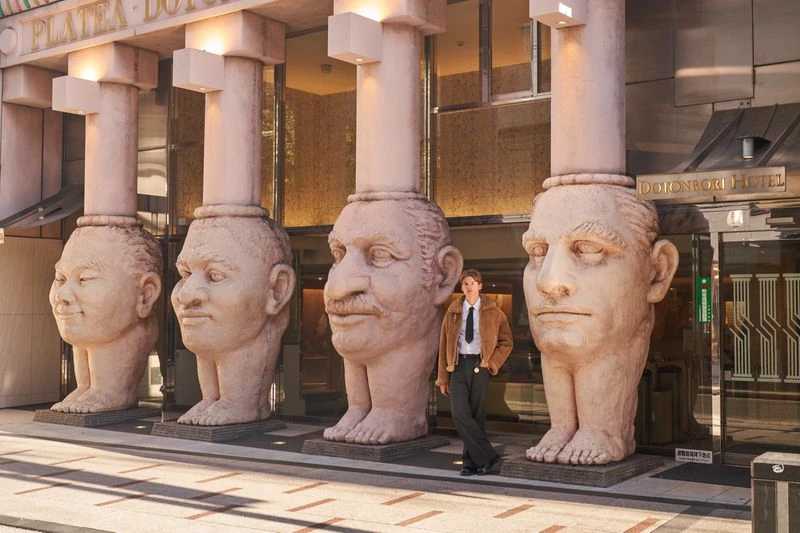 Model standing next to giant stone head columns outside Dotonbori Hotel for ODDA Magazine editorial by Regine David.