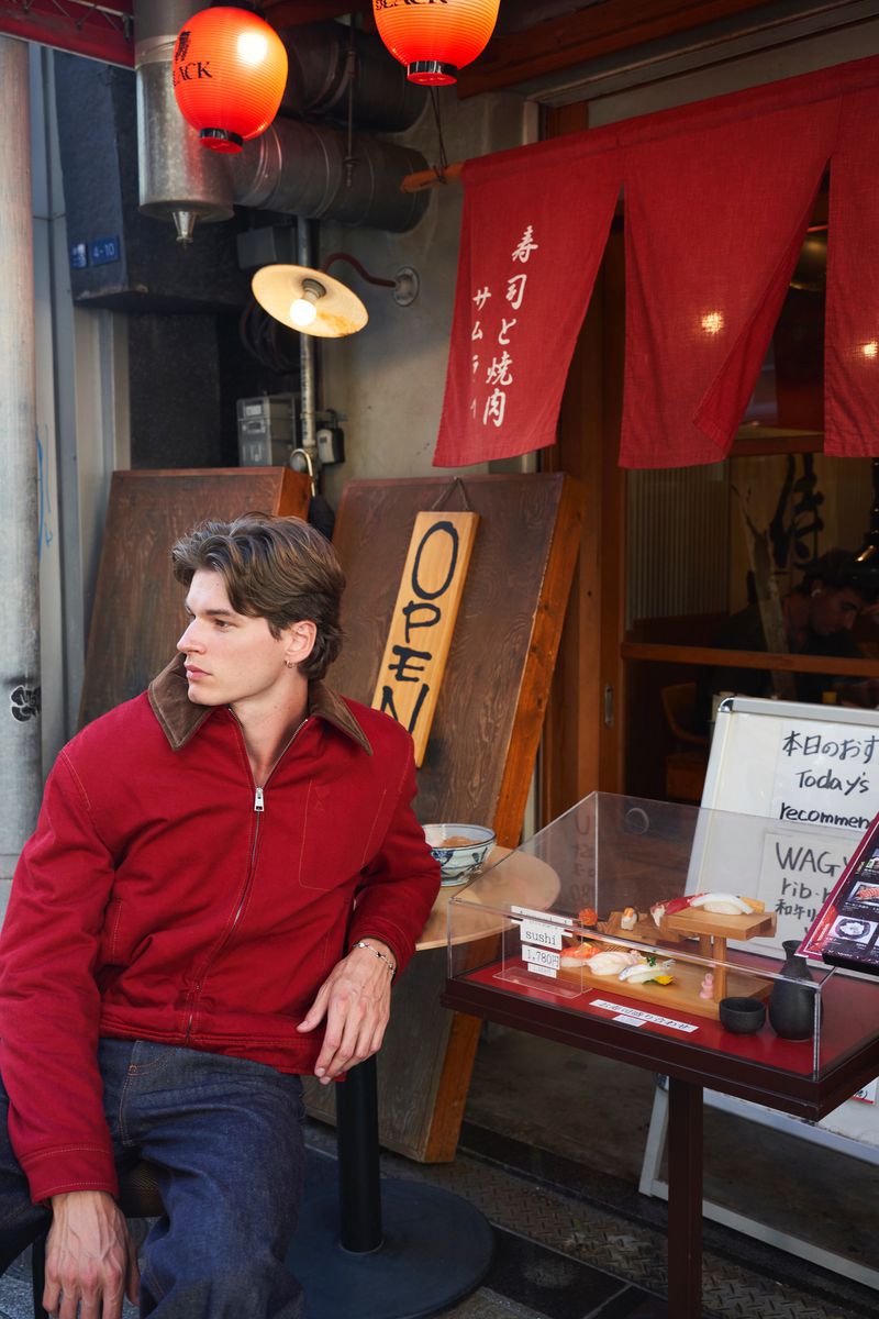 Model wearing a red jacket and denim jeans seated outside a restaurant with red lanterns and display case