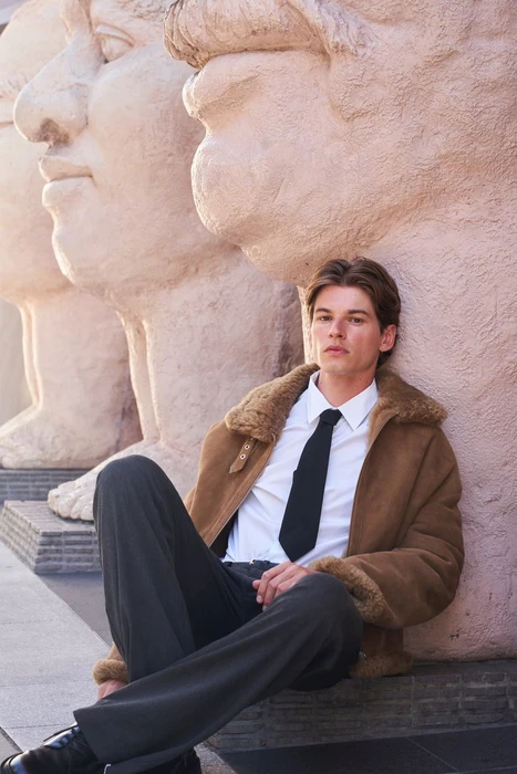 Model Jos Schenk in a brown shearling jacket and black tie leaning against giant stone statues for ODDA Magazine