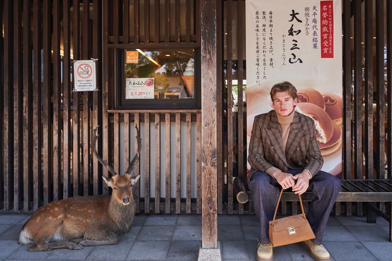 Model wearing a plaid double-breasted blazer and dark trousers seated on a wooden bench beside a resting deer in front of a shopfront.