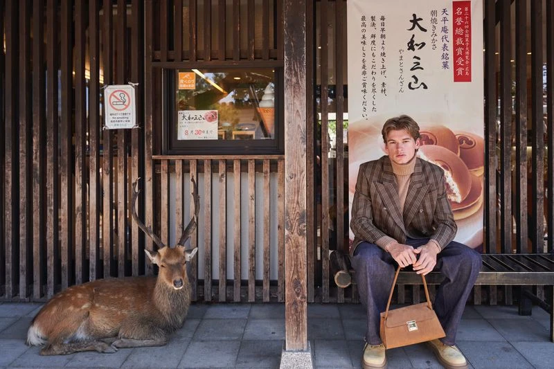 Model wearing a plaid double-breasted blazer and dark trousers seated on a wooden bench beside a resting deer in front of a shopfront.
