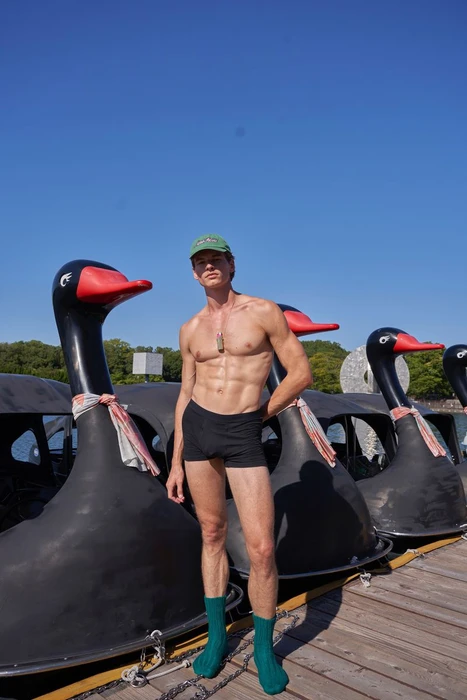 Shirtless male model wearing black underwear, green cap, and green socks standing on a wooden dock next to large swan paddle boats.
