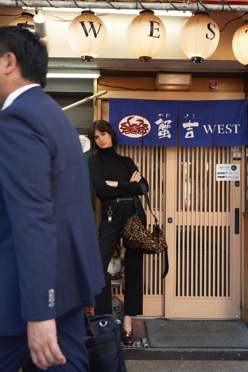 Model standing with arms crossed wearing a black turtleneck and trousers holding a leopard print bag outside a restaurant storefront