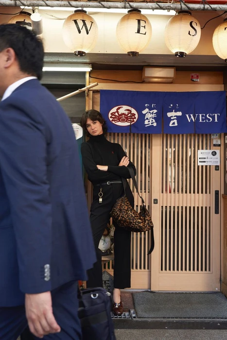 Model standing with arms crossed wearing a black turtleneck and trousers holding a leopard print bag outside a restaurant storefront