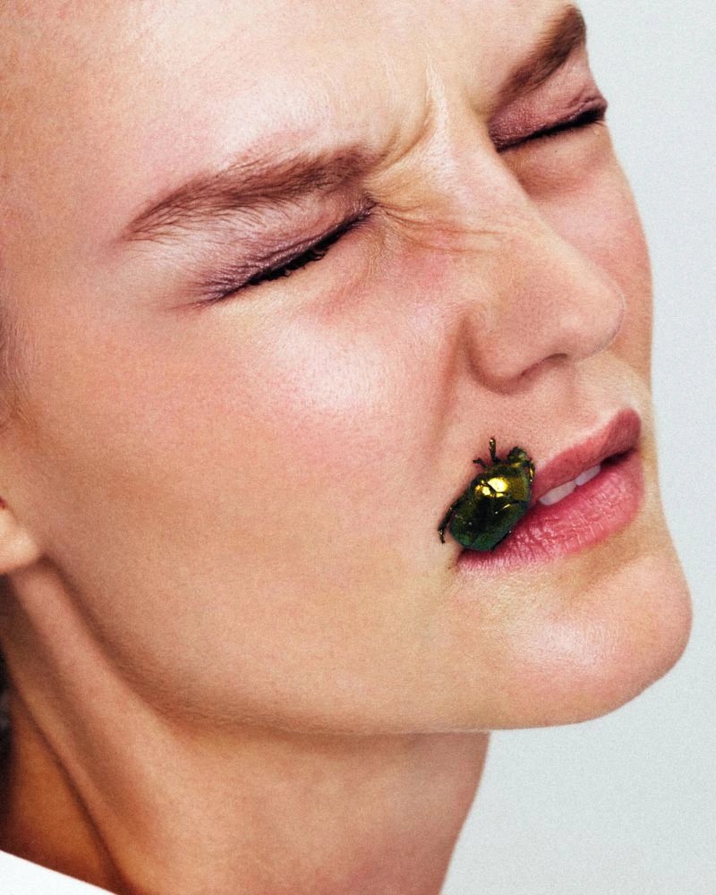 Close-up portrait of a woman with a shimmering green beetle on her lips against a neutral background.