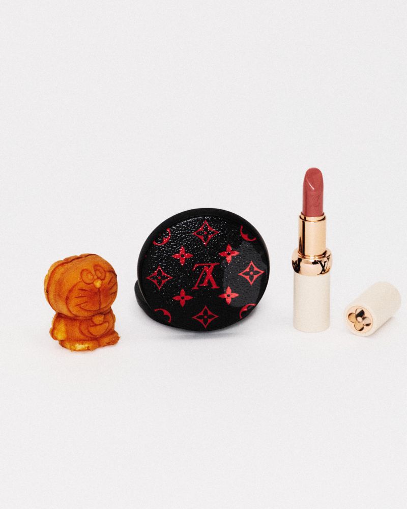 A small toy figure, a compact case with red monogram patterns, and an open lipstick tube stand against a white seamless background.