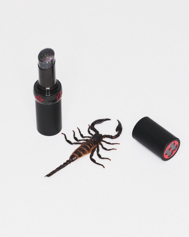 An open black lipstick tube with speckled product alongside its cap and a real dark scorpion on a white seamless background.
