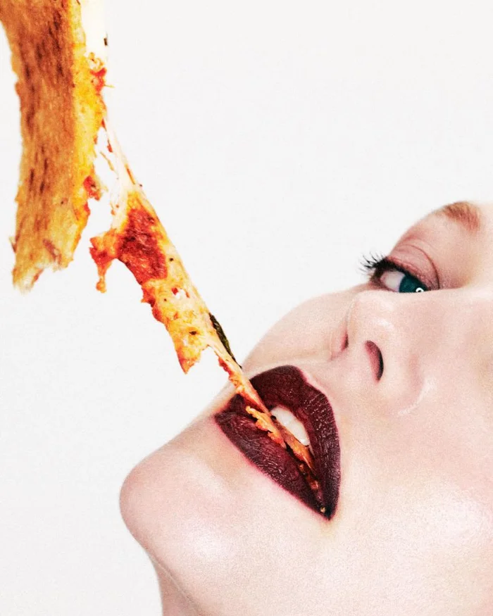 Close-up of a model eating a slice of pizza with melting cheese stretching down to dark red lips against a white background.