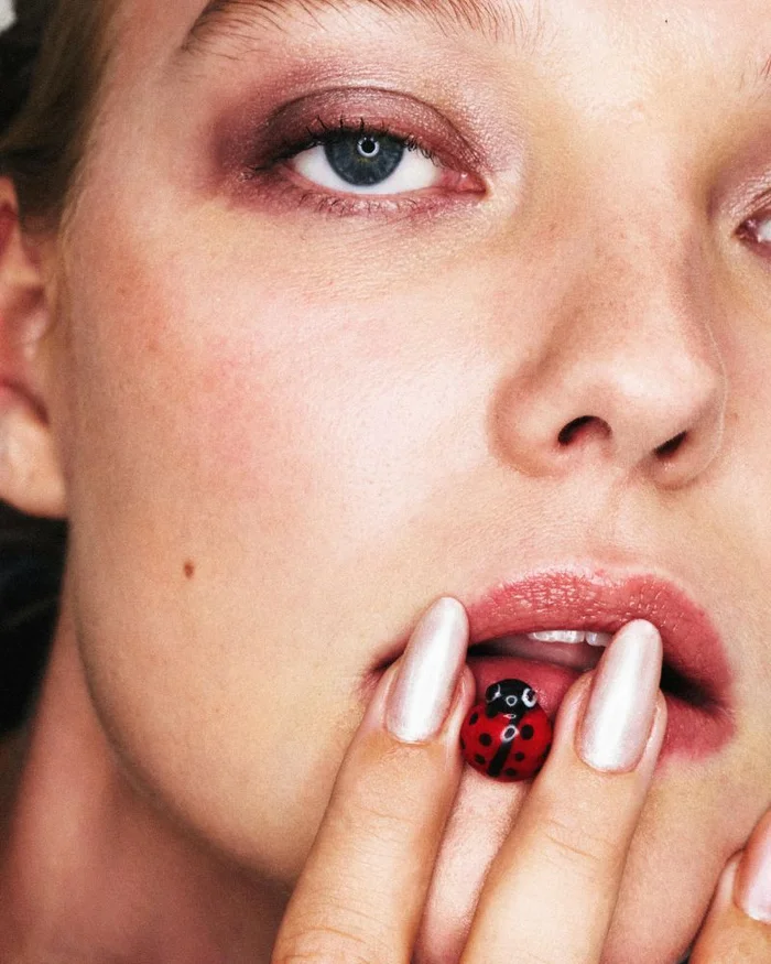 Close-up portrait of model Roos Abels with pink eyeshadow holding a small ladybug figurine against her lips for ODDA Magazine.