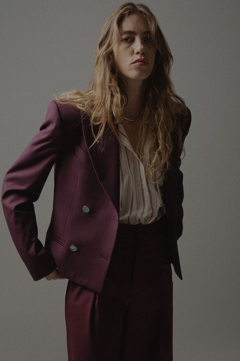 A young woman with long, wavy brown hair poses in a burgundy double-breasted blazer, striped shirt, and matching pants against a grey background.