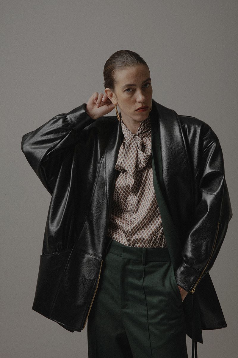 Adult woman in stylish black faux-leather coat, patterned top, and green pants poses indoors against a neutral backdrop in a fashion shot.