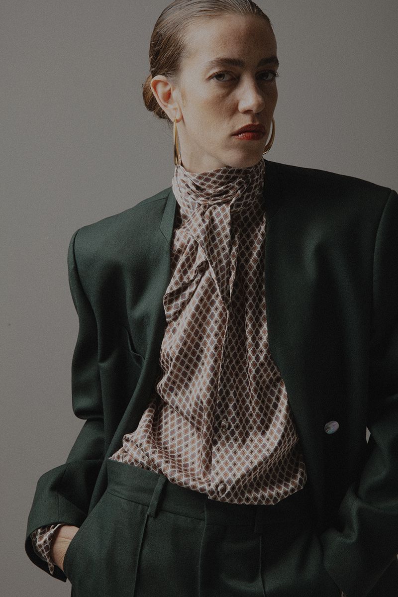 A stylish woman in a green suit and patterned blouse poses in a studio portrait, showcasing fashion and elegance with a neutral backdrop.