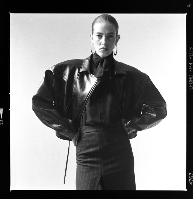 A model in a black leather jacket and pinstripe skirt poses in a studio setting. The photo is in black and white and has a film aesthetic.