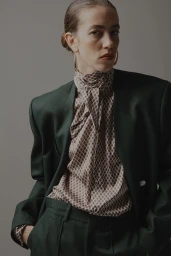 Model wearing a dark green blazer over a patterned silk high-neck blouse against a neutral grey background