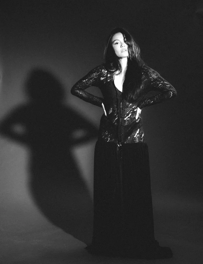 Woman in black lace top and long skirt standing against a plain background with a prominent shadow cast on the wall.