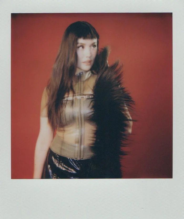 Agnez Mo in a sheer top and black feathered garment against a red background in a polaroid frame