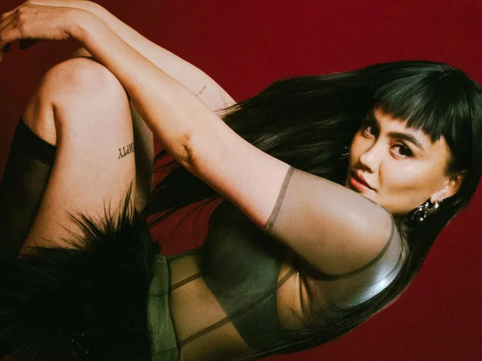 Agnez Mo with dark hair and bangs in a sheer top posing against a deep red background for Schon Magazine