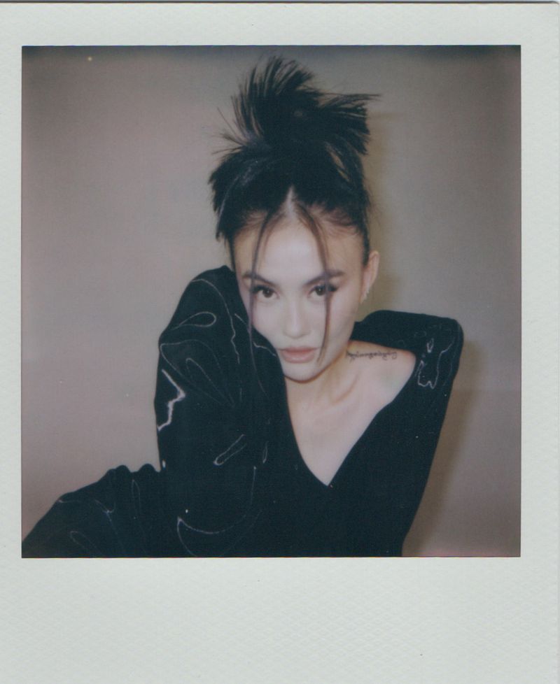 Portrait of Agnez Mo with a dramatic updo and face-framing strands wearing a dark top against a plain backdrop.
