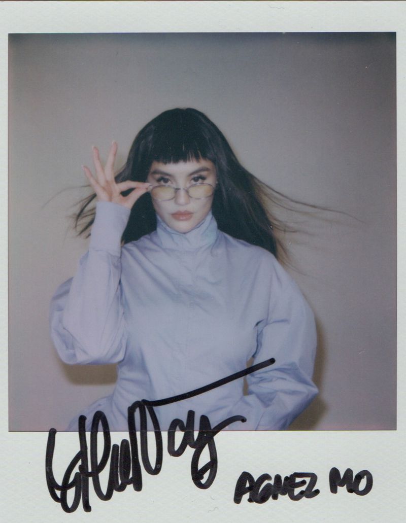 Polaroid photograph of Agnez Mo wearing a light purple turtleneck and wire-rimmed glasses against a neutral wall.