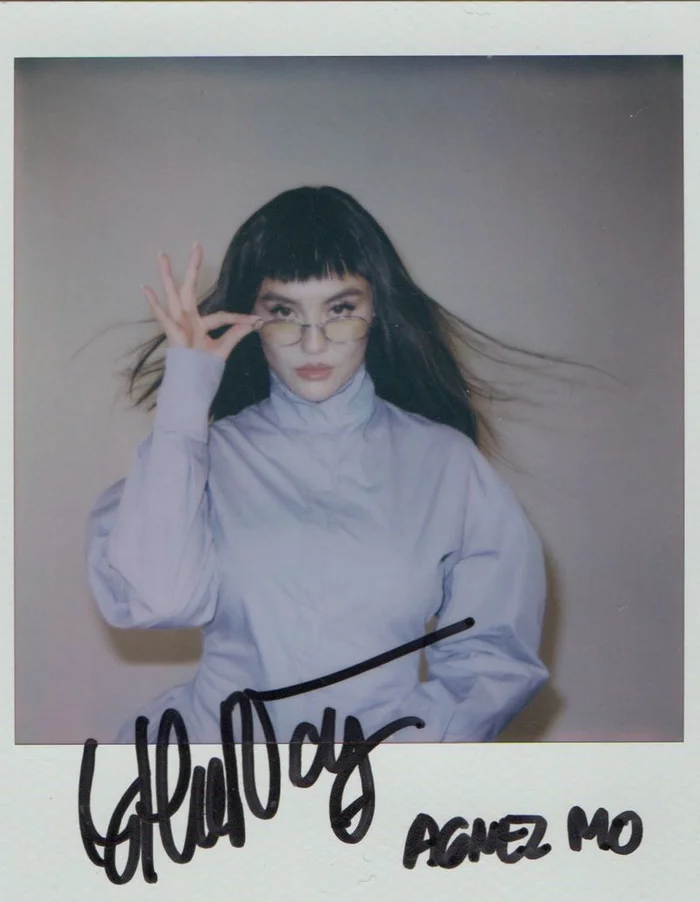 Polaroid photograph of Agnez Mo wearing a light purple turtleneck and wire-rimmed glasses against a neutral wall.