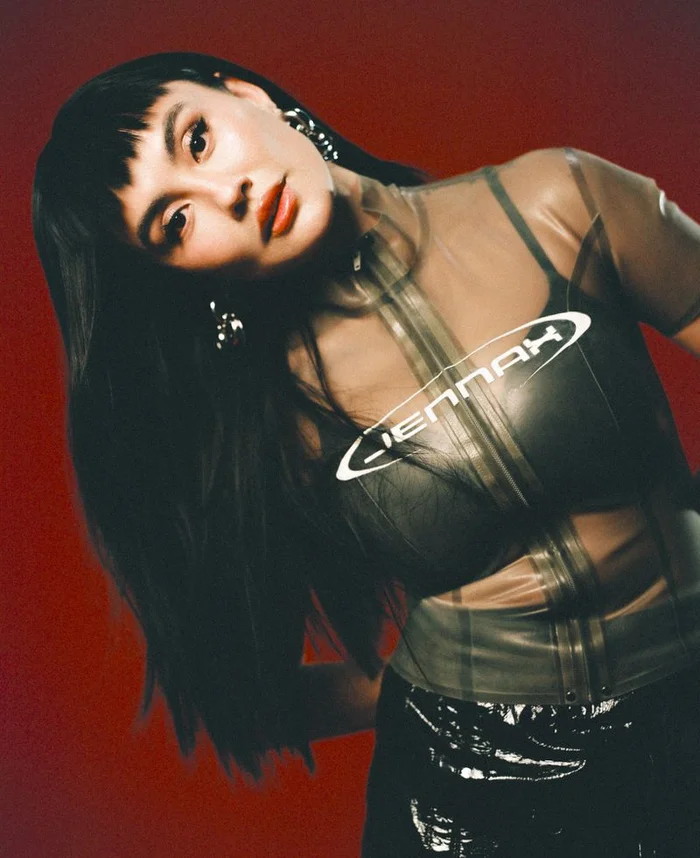 Portrait of Agnez Mo with dark hair and bangs wearing a sheer dark vinyl top against a red studio backdrop