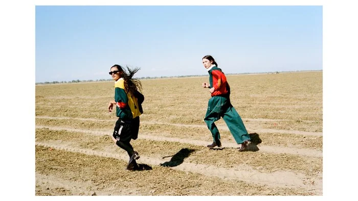 Two models running across a dry agricultural field under a clear blue sky for an Exhibition Magazine fashion editorial.