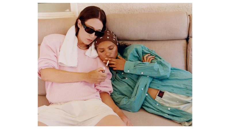 Two models relaxing on a beige sofa, one holding a lighter while the other smokes, for Exhibition Magazine.