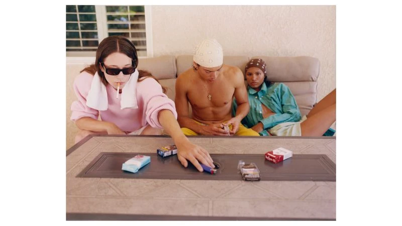 Three models gathered around an outdoor table with cigarette packs and a lighter, featured in Exhibition Magazine