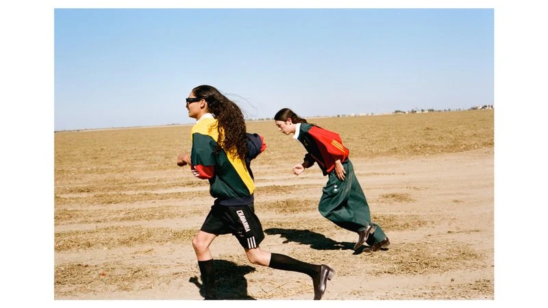 Two models in sportswear running across a dry dirt field under a clear blue sky for Exhibition Magazine.