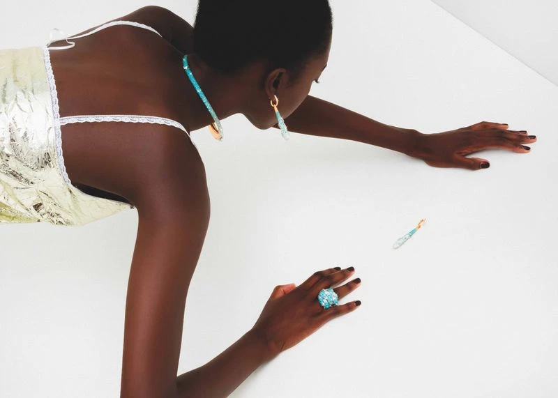 Model with dark hair wearing a light metallic dress and turquoise jewelry reaching forward against white background